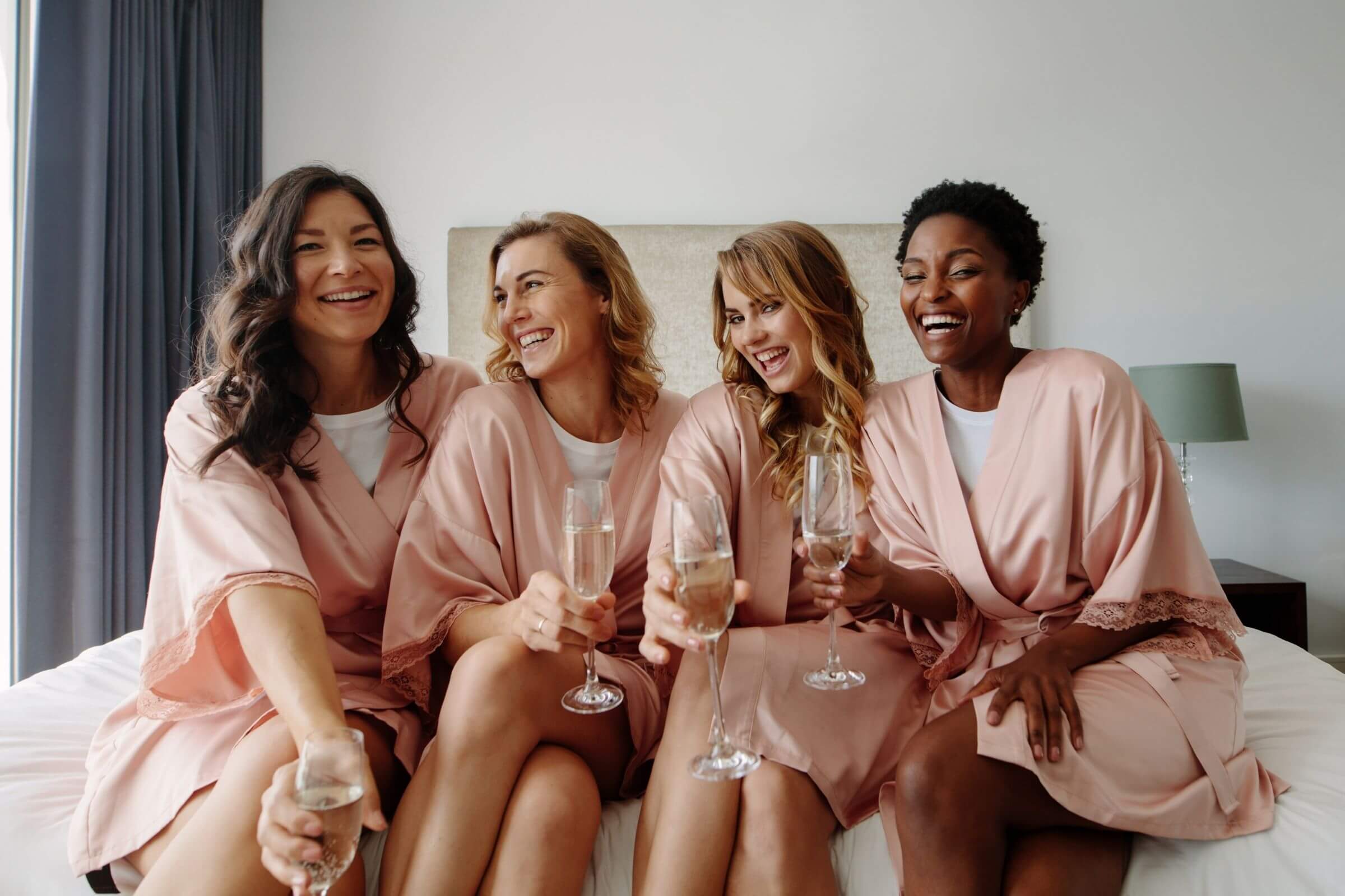 why to send bridesmaid boxes - four women smiling sitting on a bed with champagne